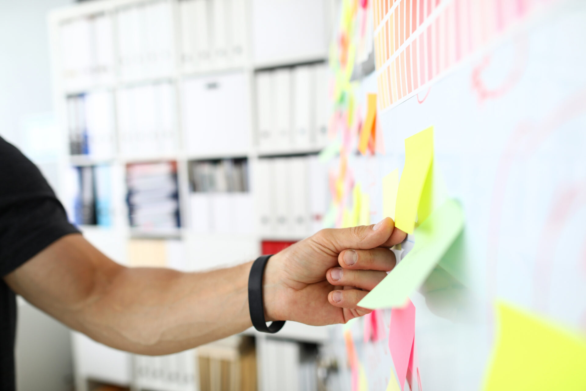 Hand mit schwarzem Armband klebt bunte Haftnotizen an eine weiße Wand mit weiteren Haftnotizen