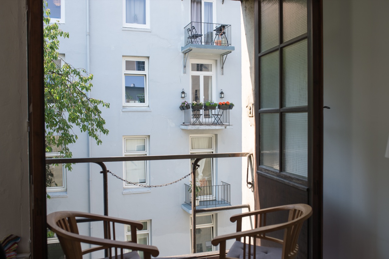 Blick aus einem Raum auf einen Balkon mit zwei Holzstühlen und einem Geländer vor einem gegenüberliegenden Wohnhaus mit Balkonen und Fenstern
