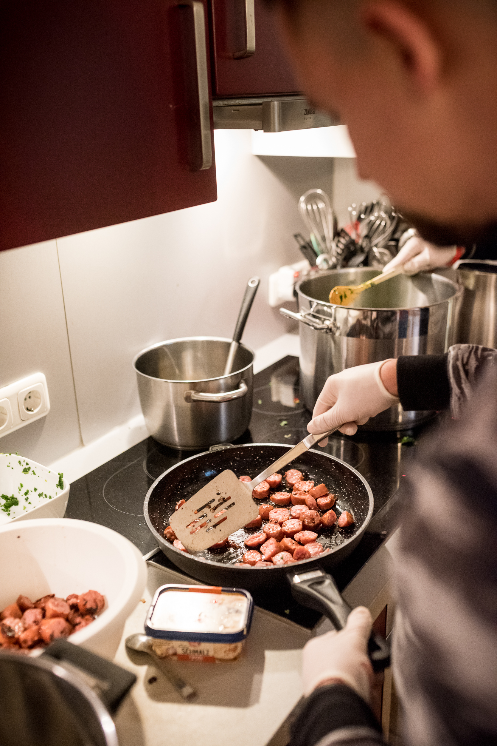Person brät Wurstscheiben in einer Pfanne neben mehreren Edelstahltöpfen und Schüsseln auf einer Küchenarbeitsfläche