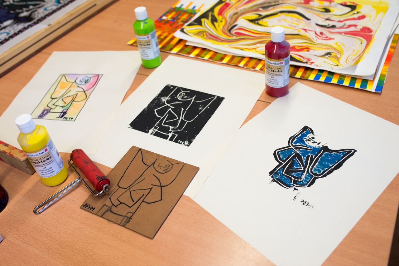 Mehrere Drucke mit stilisierten Figuren in verschiedenen Farben liegen auf einem Tisch neben Flaschen mit Farbe und einer Farbwalze