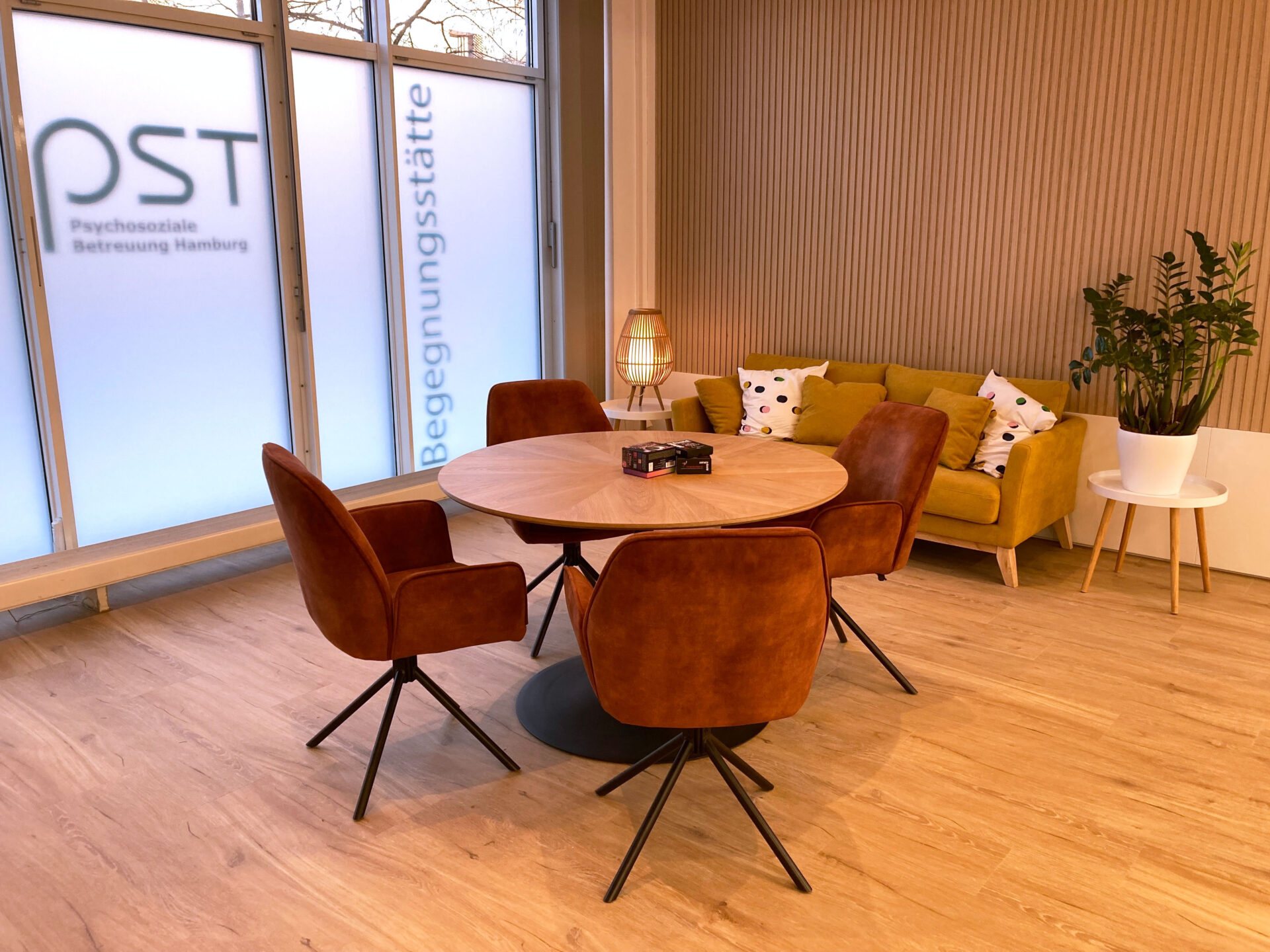 Runder Holztisch mit vier braunen Stühlen vor großen Fenstern neben gelbem Sofa mit Kissen und Pflanzen in heller Begegnungsstätte